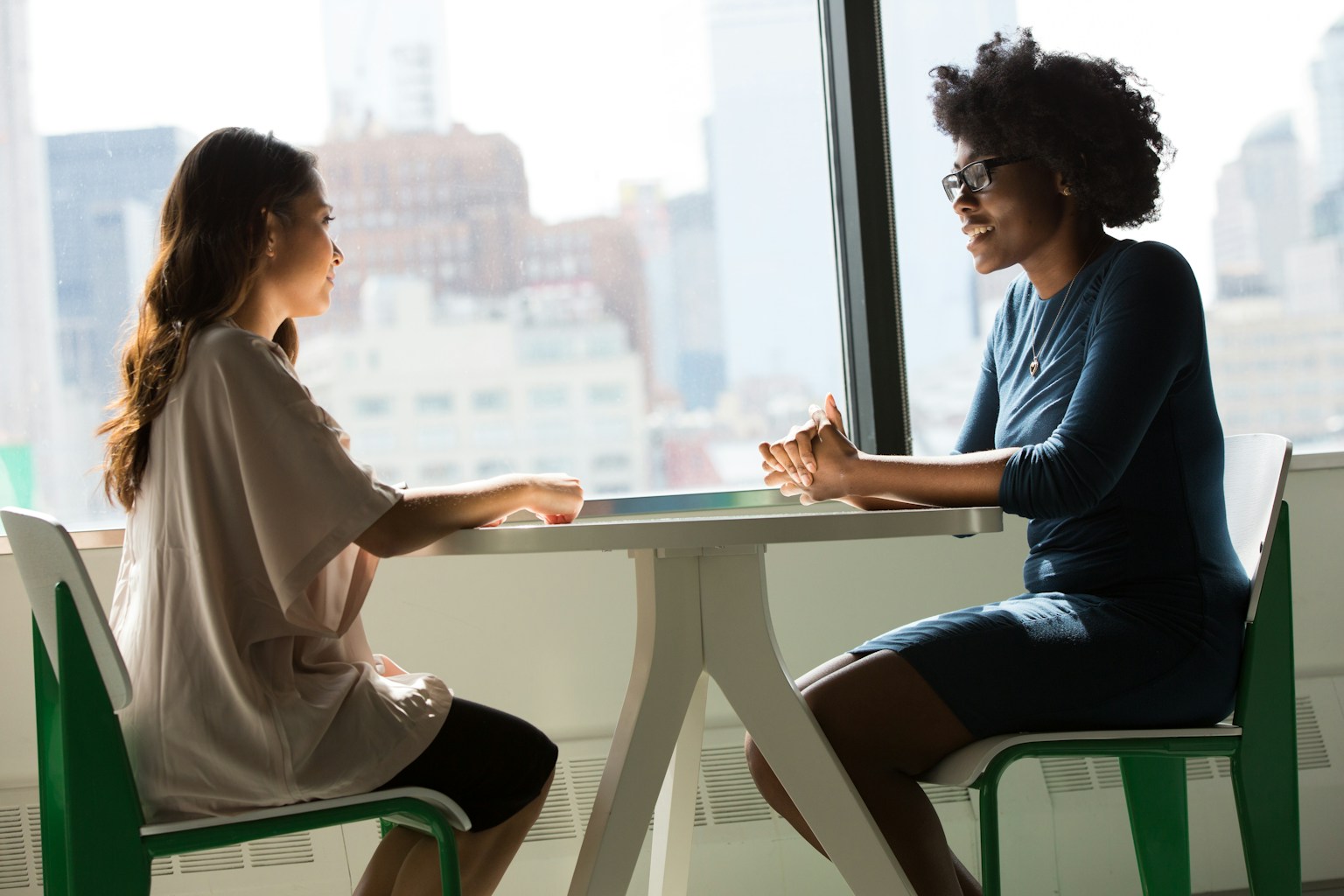 Two women in conversation across a table, one listening intently, city skyline behind them